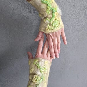 May include: A pair of beige felted wool fingerless gloves featuring raised, textured floral patterns in shades of light green and yellow. The fabric has a soft, fibrous appearance with scalloped edges at the wrist and hand openings.