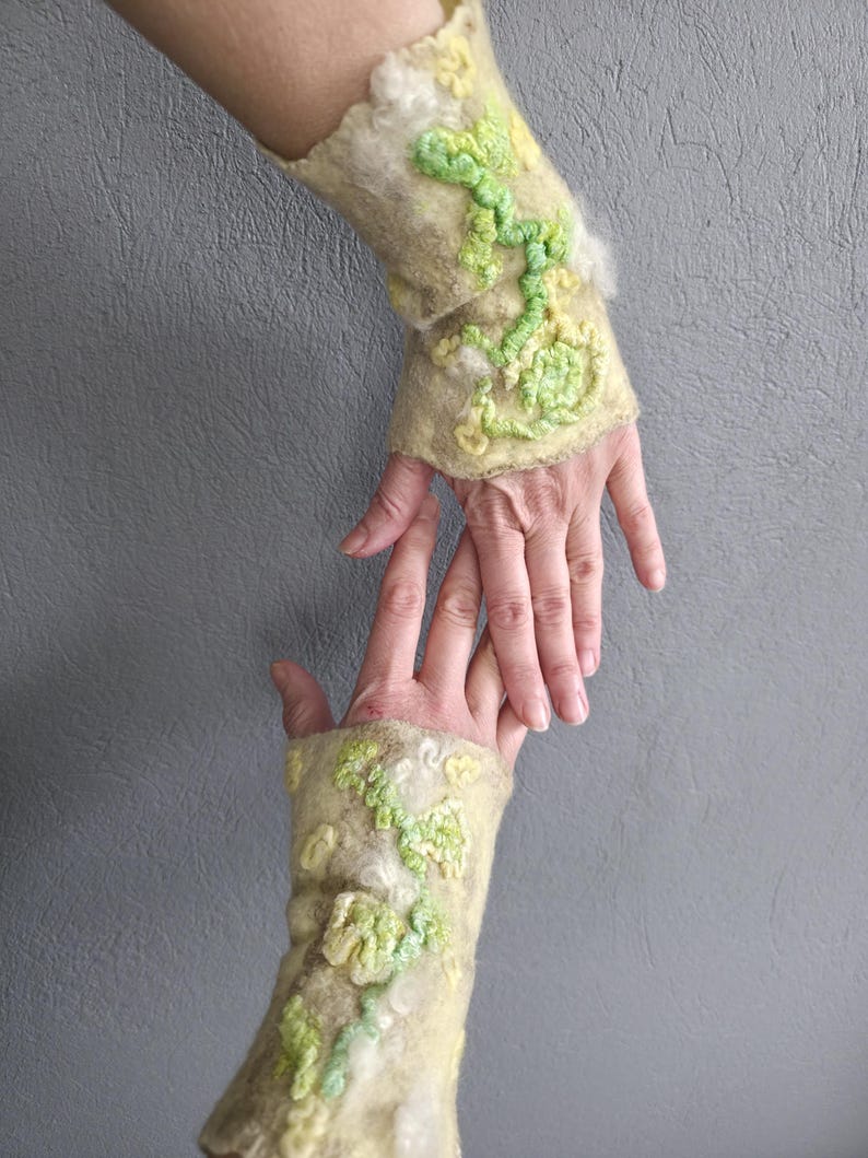 May include: A pair of beige felted wool fingerless gloves featuring raised, textured floral patterns in shades of light green and yellow. The fabric has a soft, fibrous appearance with scalloped edges at the wrist and hand openings.