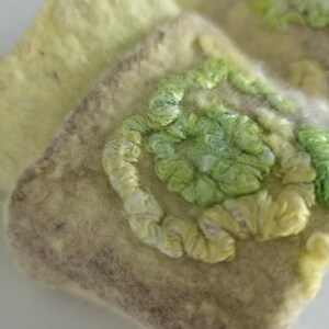 May include: A square-shaped felted textile piece in shades of pale yellow and beige, featuring a raised, swirling embroidery pattern in vibrant lime and light green thread. The texture is soft and fibrous, with a distinct contrast between the base.