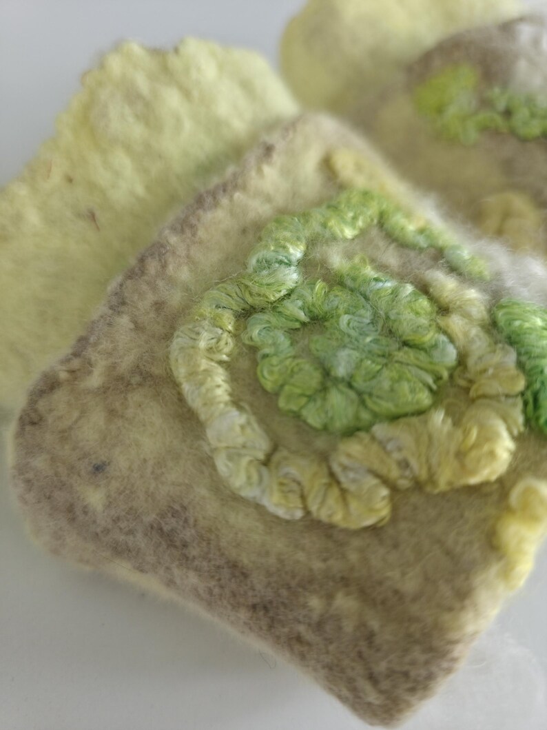 May include: A square-shaped felted textile piece in shades of pale yellow and beige, featuring a raised, swirling embroidery pattern in vibrant lime and light green thread. The texture is soft and fibrous, with a distinct contrast between the base.