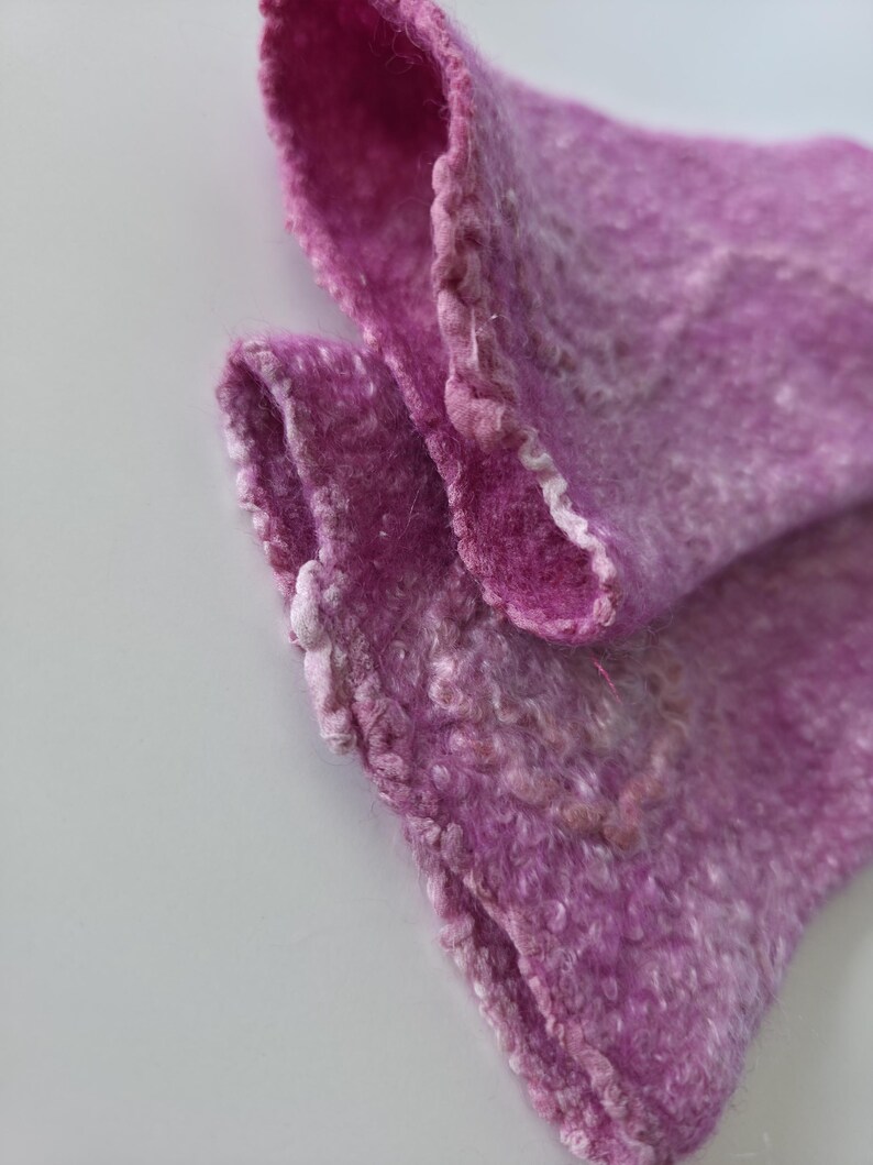 May include: Close-up of a felted textile in shades of purple. The fabric has a textured, slightly uneven surface with visible fibres. The edges are softly defined, with a lighter, almost white, border. The textile is folded, revealing its layered construction.
