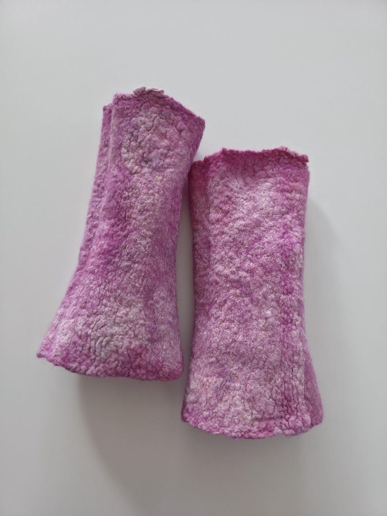 May include: A pair of magenta-coloured arm warmers made from a textured, felted material. The arm warmers have a flared shape at the wrist and a slightly ruffled edge. The background is plain white. The arm warmers are handmade.