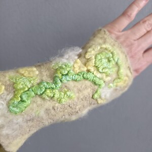 May include: A beige felted wrist warmer featuring raised, textured embroidery in shades of lime green, pale yellow, and white. The surface has a fuzzy, fibrous texture with swirling, organic patterns created by thick, looped yarn stitching.