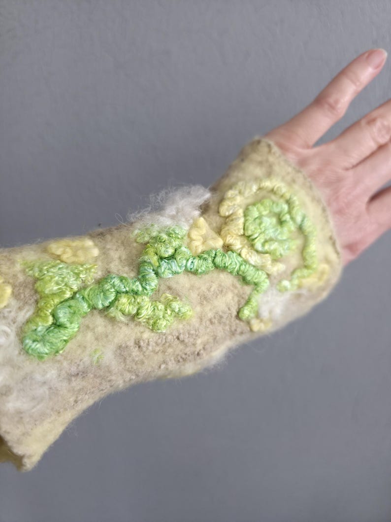 May include: A beige felted wrist warmer featuring raised, textured embroidery in shades of lime green, pale yellow, and white. The surface has a fuzzy, fibrous texture with swirling, organic patterns created by thick, looped yarn stitching.