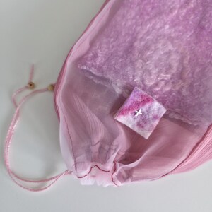 May include: A pink mesh drawstring bag with a light pink lining, containing a soft, textured item. A small, square, felted item with a white cross is on top of the bag. The bag is tied with a pink cord and wooden bead.