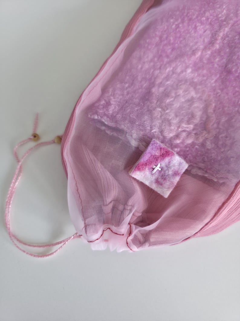 May include: A pink mesh drawstring bag with a light pink lining, containing a soft, textured item. A small, square, felted item with a white cross is on top of the bag. The bag is tied with a pink cord and wooden bead.