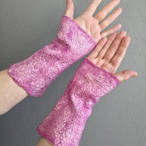 May include: A pair of magenta-coloured, felted wool wrist warmers. The warmers are a vibrant pink with a textured, mottled appearance. The open-finger design allows for dexterity while keeping the wrists warm. The background is a solid grey.