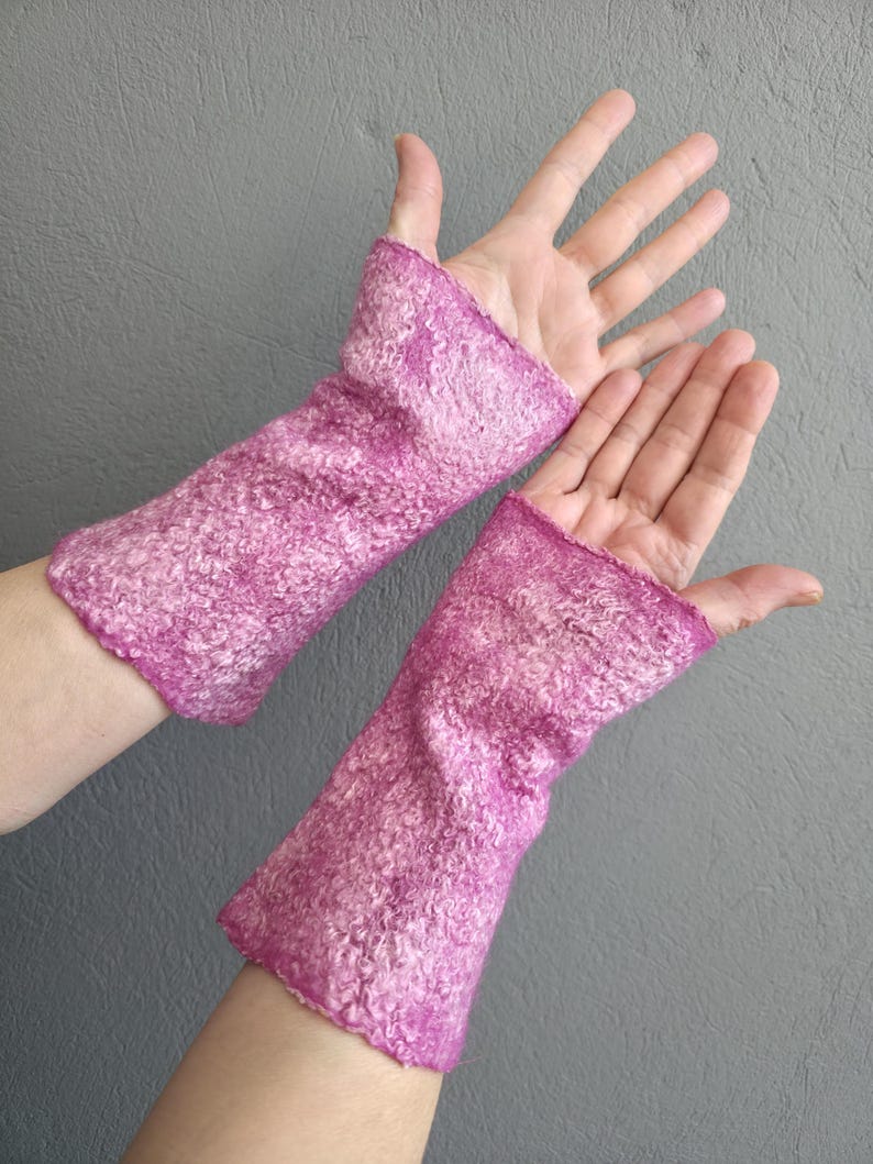 May include: A pair of magenta-coloured, felted wool wrist warmers. The warmers are a vibrant pink with a textured, mottled appearance. The open-finger design allows for dexterity while keeping the wrists warm. The background is a solid grey.