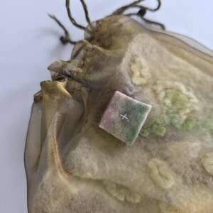 May include: A sheer olive-gold organza drawstring pouch containing a textured, off-white felted textile with light green embroidered floral details. A small, square, multi-colored felt patch with a single cross-stitch is attached to the front.