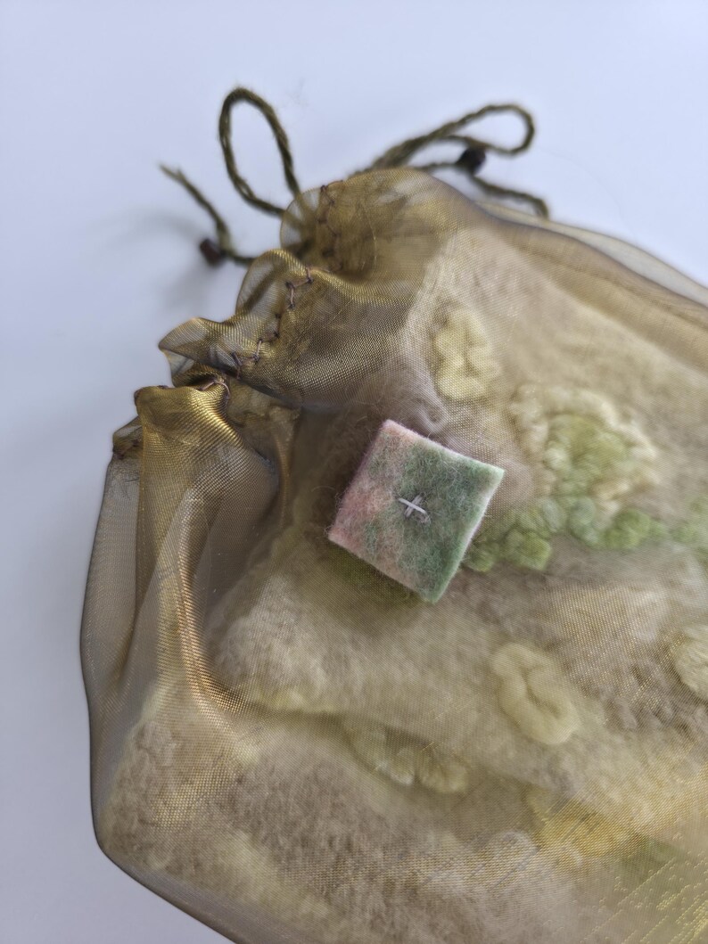 May include: A sheer olive-gold organza drawstring pouch containing a textured, off-white felted textile with light green embroidered floral details. A small, square, multi-colored felt patch with a single cross-stitch is attached to the front.