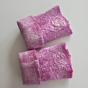 May include: A pair of handmade, felted wrist warmers in shades of pink and white. The textured wool cuffs are folded over, and one warmer features a small, lighter-coloured floral design. The background is a plain white surface.