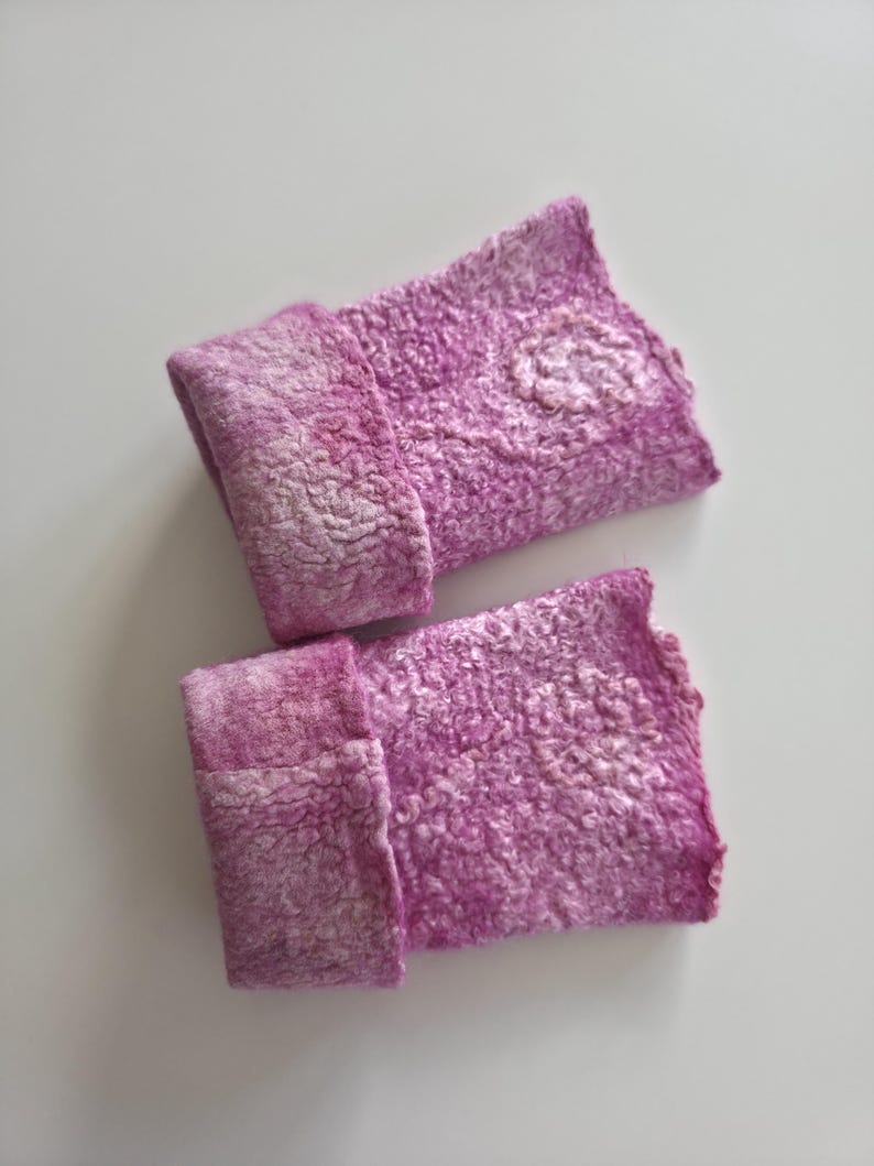 May include: A pair of handmade, felted wrist warmers in shades of pink and white. The textured wool cuffs are folded over, and one warmer features a small, lighter-coloured floral design. The background is a plain white surface.