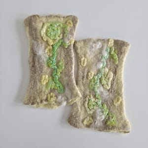 May include: A pair of beige felted wool fingerless gloves featuring a textured floral design in shades of light green and yellow. The surface has a soft, fibrous appearance with raised, sculpted flower and vine motifs across the front.