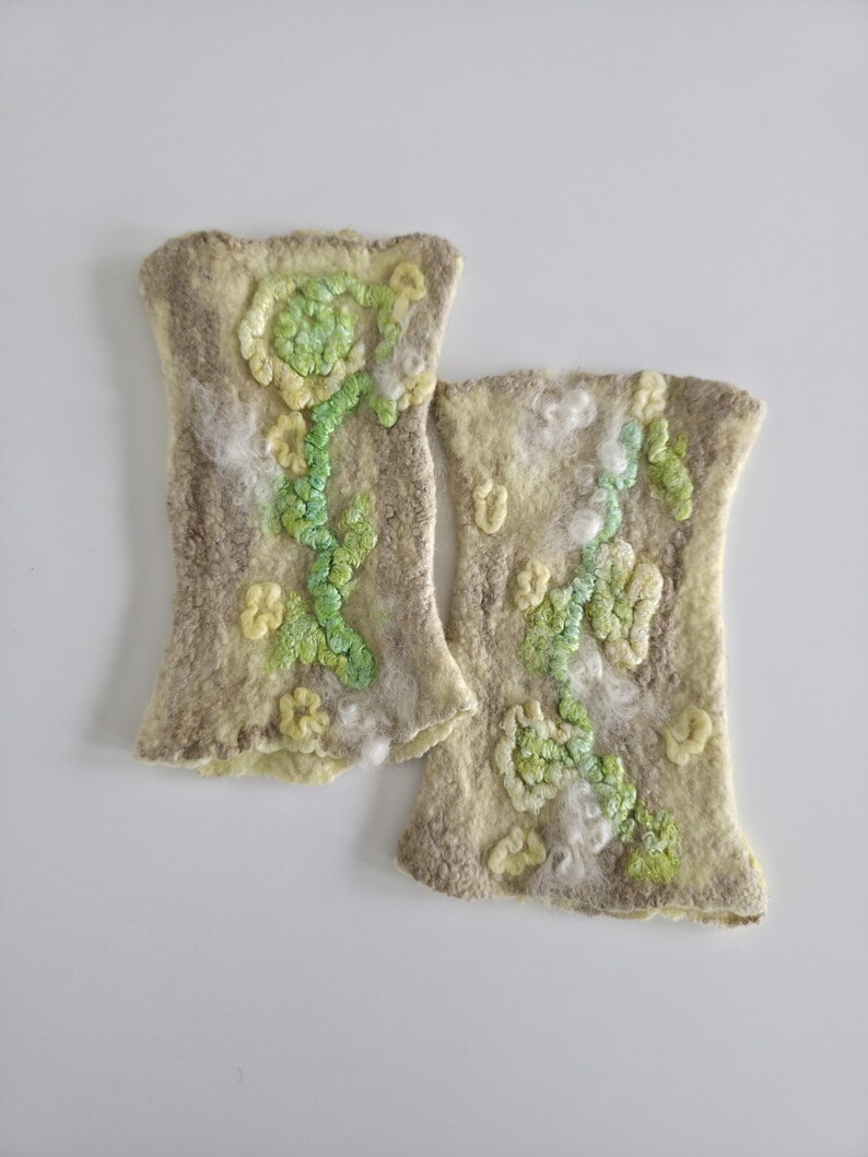 May include: A pair of beige felted wool fingerless gloves featuring a textured floral design in shades of light green and yellow. The surface has a soft, fibrous appearance with raised, sculpted flower and vine motifs across the front.