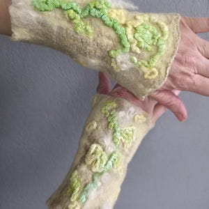 May include: A pair of light beige felted wrist warmers featuring raised, textured floral embroidery in shades of lime green, pale yellow, and white. The surface has a soft, fibrous texture with organic, swirling patterns and dimensional details.