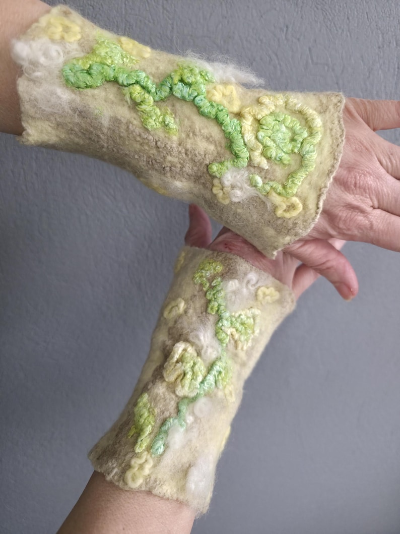 May include: A pair of light beige felted wrist warmers featuring raised, textured floral embroidery in shades of lime green, pale yellow, and white. The surface has a soft, fibrous texture with organic, swirling patterns and dimensional details.