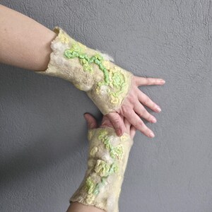 May include: A pair of beige felted wool fingerless gloves featuring textured, raised floral patterns in shades of light green and pale yellow. The fabric has a soft, fibrous appearance with an uneven, organic edge at the wrist and hand openings.