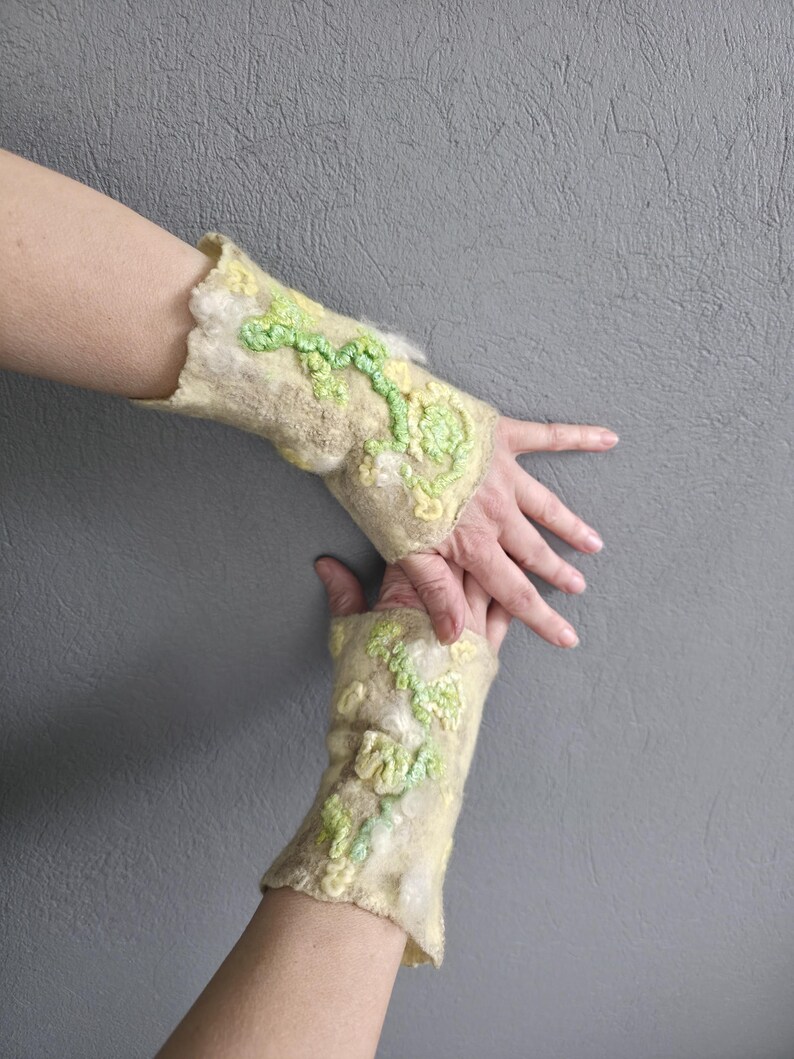 May include: A pair of beige felted wool fingerless gloves featuring textured, raised floral patterns in shades of light green and pale yellow. The fabric has a soft, fibrous appearance with an uneven, organic edge at the wrist and hand openings.