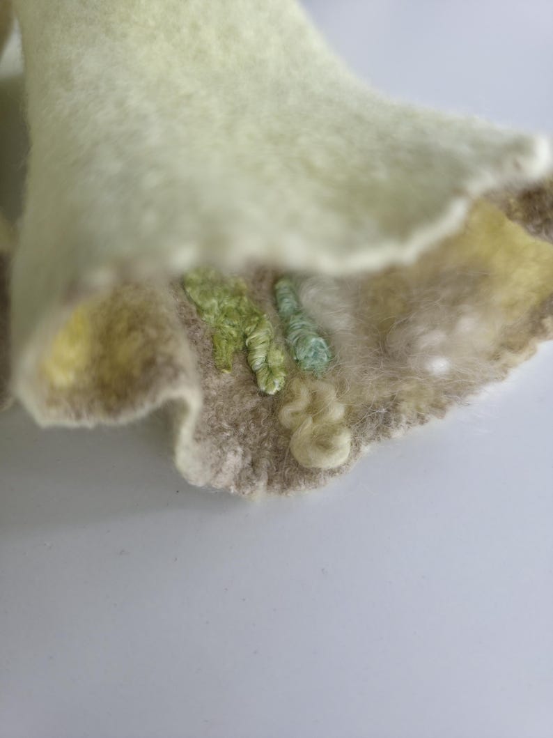 May include: A piece of light green felted fabric with a wavy, scalloped edge. The surface features textured embroidery in shades of light green, mint, and cream, with small, raised loops and tufts of fiber creating a tactile, dimensional pattern.