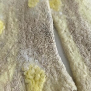 May include: A pair of felted wool fingerless gloves featuring a textured beige base with irregular, raised pale yellow spots. The material has a dense, matted appearance with visible fiber loops and a soft, uneven surface throughout the fabric.