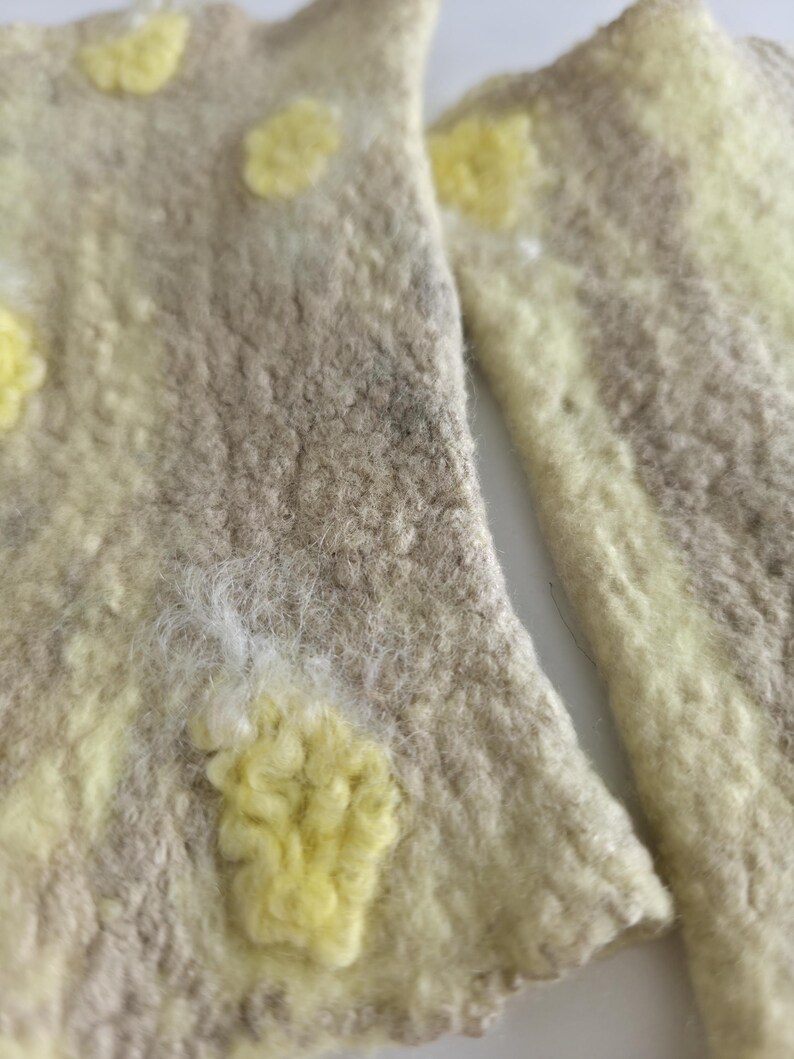 May include: A pair of felted wool fingerless gloves featuring a textured beige base with irregular, raised pale yellow spots. The material has a dense, matted appearance with visible fiber loops and a soft, uneven surface throughout the fabric.