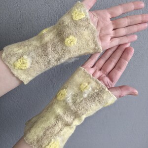 May include: A pair of fingerless gloves crafted from felted wool in a pale beige and cream color palette. The gloves feature a textured surface with raised, circular yellow floral-like embellishments scattered across the fabric.