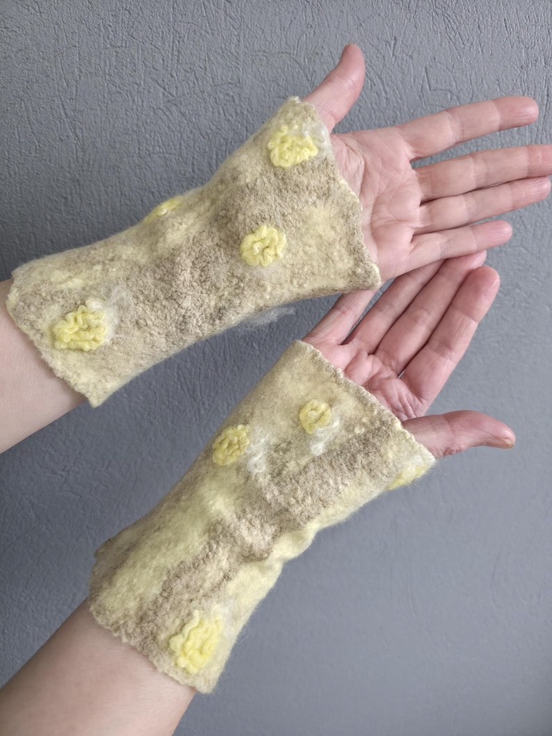 May include: A pair of fingerless gloves crafted from felted wool in a pale beige and cream color palette. The gloves feature a textured surface with raised, circular yellow floral-like embellishments scattered across the fabric.