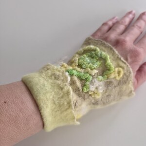 May include: A light yellow and beige felted wrist warmer featuring a textured, raised floral embroidery design in shades of lime green, pale yellow, and white. The accessory has a soft, fuzzy surface texture and a wide, folded cuff edge.