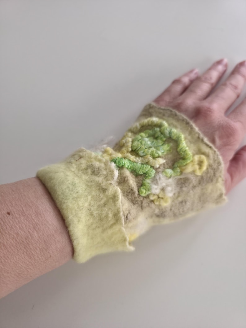 May include: A light yellow and beige felted wrist warmer featuring a textured, raised floral embroidery design in shades of lime green, pale yellow, and white. The accessory has a soft, fuzzy surface texture and a wide, folded cuff edge.