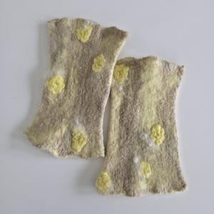May include: A pair of fingerless gloves made from felted wool in a light beige and tan color palette. The gloves feature a textured surface with scattered, raised yellow floral-like embellishments and a soft, uneven edge at the openings.