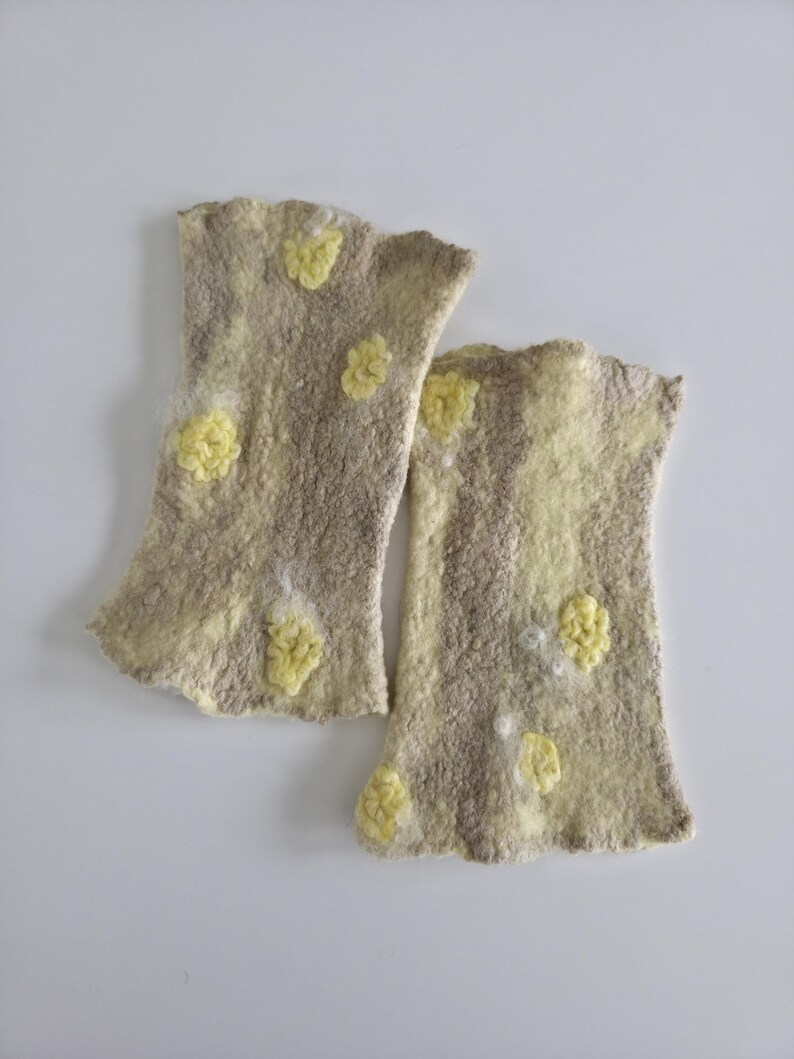 May include: A pair of fingerless gloves made from felted wool in a light beige and tan color palette. The gloves feature a textured surface with scattered, raised yellow floral-like embellishments and a soft, uneven edge at the openings.