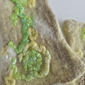 May include: A pair of beige felted wool cuffs featuring textured embroidery in shades of light green, lime, and pale yellow. The surface displays a swirling, organic pattern of raised yarn loops and soft, fuzzy white fiber accents.