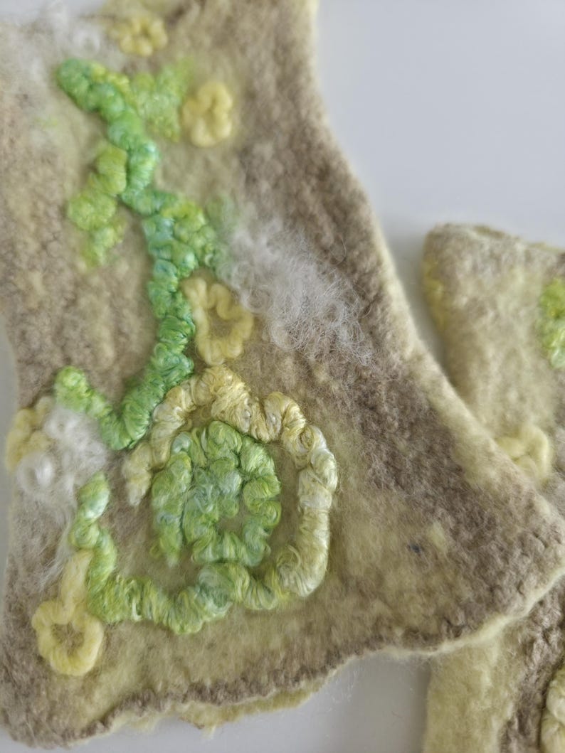 May include: A pair of beige felted wool cuffs featuring textured embroidery in shades of light green, lime, and pale yellow. The surface displays a swirling, organic pattern of raised yarn loops and soft, fuzzy white fiber accents.