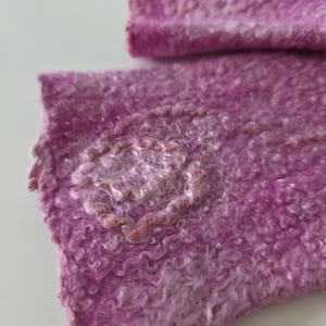 May include: Close-up of a textured, handmade textile in shades of pink and purple. The fabric has a curly, wool-like appearance with a lighter, circular design element. The overall aesthetic suggests a soft, handcrafted item.