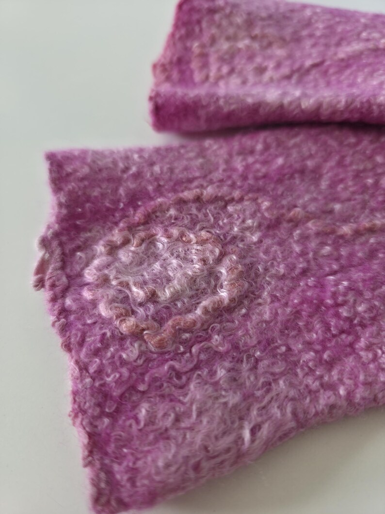 May include: Close-up of a textured, handmade textile in shades of pink and purple. The fabric has a curly, wool-like appearance with a lighter, circular design element. The overall aesthetic suggests a soft, handcrafted item.