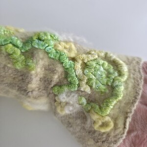 May include: A beige felted wrist warmer featuring raised, textured embroidery in shades of light green, lime, and pale yellow. The surface displays a dense, looped yarn pattern that creates a three-dimensional floral or vine-like design across the fabric.