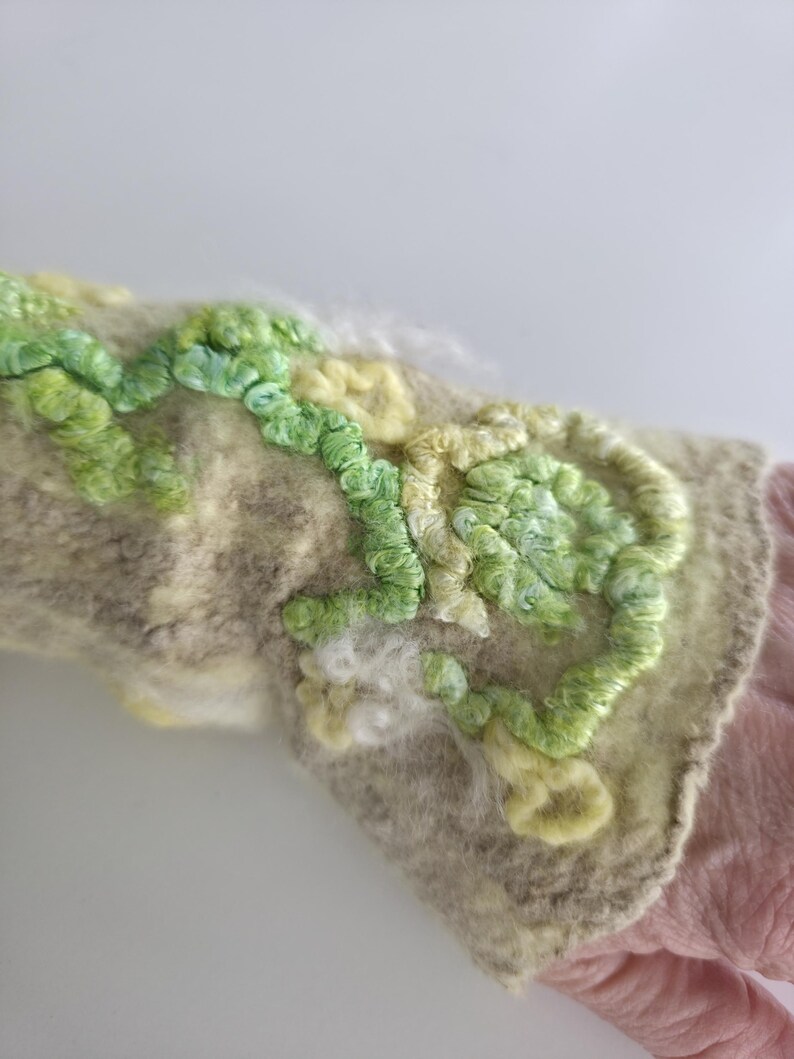 May include: A beige felted wrist warmer featuring raised, textured embroidery in shades of light green, lime, and pale yellow. The surface displays a dense, looped yarn pattern that creates a three-dimensional floral or vine-like design across the fabric.
