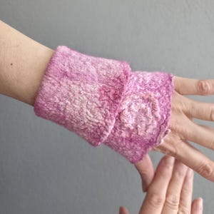 May include: Handmade pink wrist warmers. The warmers are made of a textured, felted material with a slightly ruffled edge. The colour is a blend of light and dark pink hues.