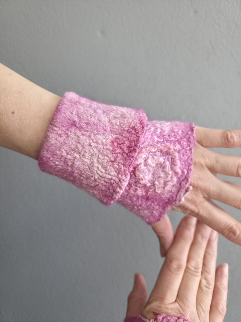 May include: Handmade pink wrist warmers. The warmers are made of a textured, felted material with a slightly ruffled edge. The colour is a blend of light and dark pink hues.