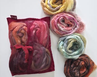 Puede incluir: Una bolsa rectangular de malla color burdeos que contiene cuatro paquetes de fibras multicolores, atada con una cinta a juego y etiquetada con una pequeña etiqueta que dice DanG. Junto a la bolsa hay cuatro paquetes circulares de fibra sueltos en tonos amarillo, rosa, verde y marrón.