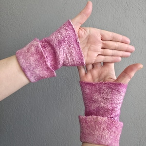 May include: A pair of pink felted wrist warmers. The warmers are a vibrant pink with a textured surface and a folded cuff. The background is a solid grey.
