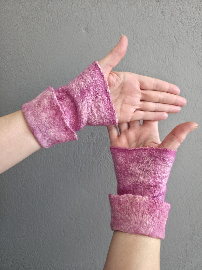 May include: A pair of pink felted wrist warmers. The warmers are a vibrant pink with a textured surface and a folded cuff. The background is a solid grey.