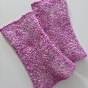 May include: Two lavender wrist warmers with a textured, fuzzy surface. The wrist warmers are rectangular in shape and made from a soft material. They are designed to keep the wrists warm. The background is a plain white surface.