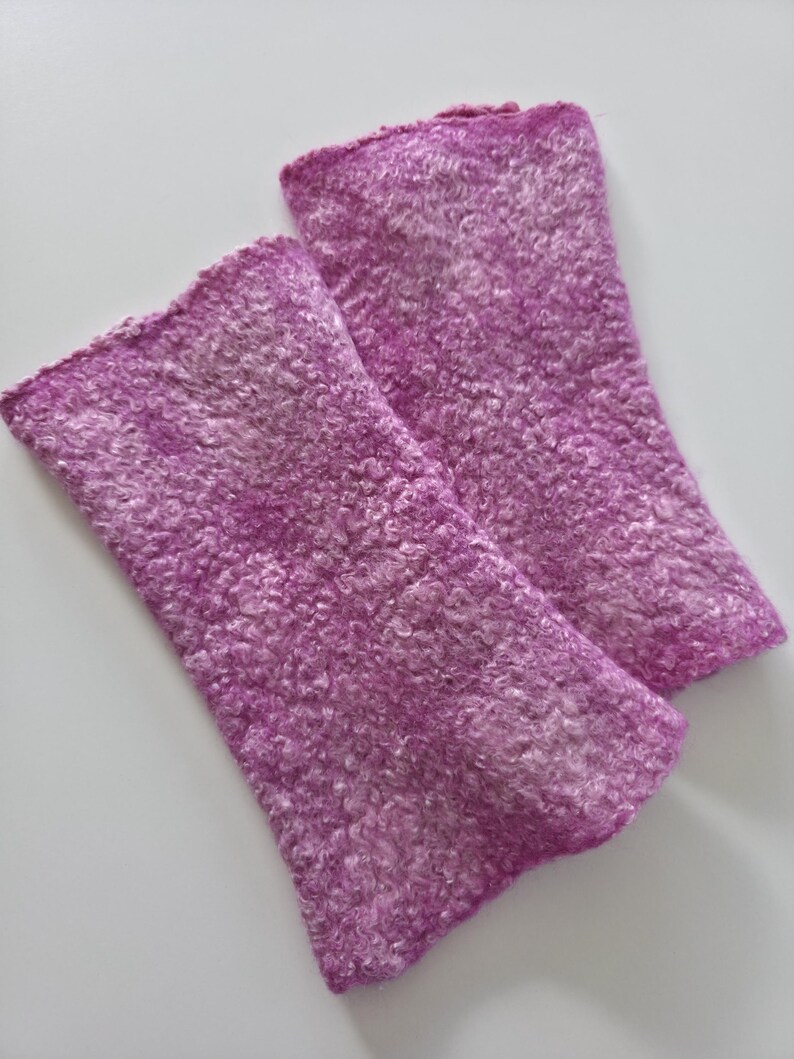 May include: Two lavender wrist warmers with a textured, fuzzy surface. The wrist warmers are rectangular in shape and made from a soft material. They are designed to keep the wrists warm. The background is a plain white surface.