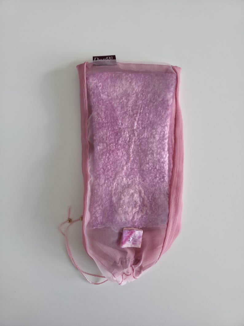 May include: A rectangular, pink, felted textile is encased in a sheer, light pink fabric pouch. The pouch has a drawstring closure and a small, square embellishment. The label at the top reads "Dante".