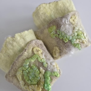 May include: Two rectangular felted wool cuffs in pale yellow and beige. Each cuff features raised, textured embroidery in shades of lime green, light green, and cream, forming swirling, organic patterns across the surface of the felted material.