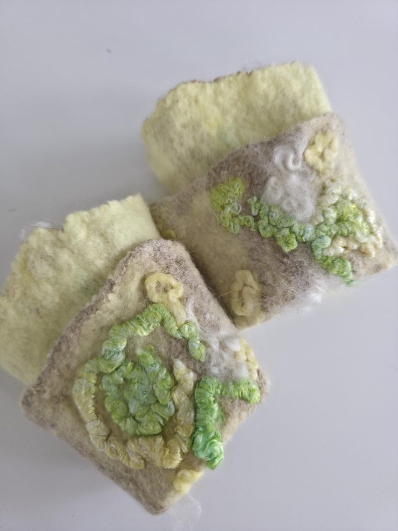 May include: Two rectangular felted wool cuffs in pale yellow and beige. Each cuff features raised, textured embroidery in shades of lime green, light green, and cream, forming swirling, organic patterns across the surface of the felted material.