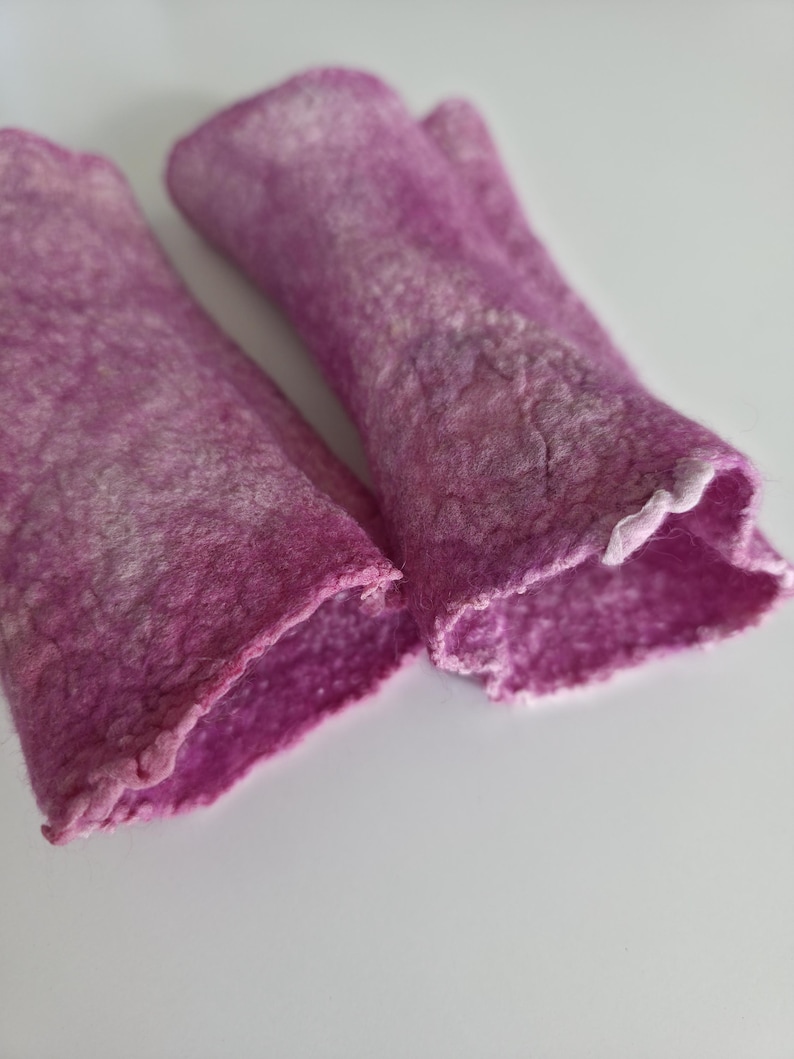 May include: A pair of magenta fingerless gloves, handmade from felted wool. The gloves have a textured finish and a ruffled edge at the wrist. They are designed to keep hands warm while allowing finger movement. The gloves are a vibrant shade of pink.