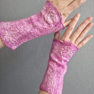 May include: A pair of pink and white felted fingerless gloves. The gloves are a vibrant pink with white flecks, and have a textured, woolly appearance. The wrist cuffs are slightly ruffled. The gloves are designed to keep hands warm while leaving fingers free.