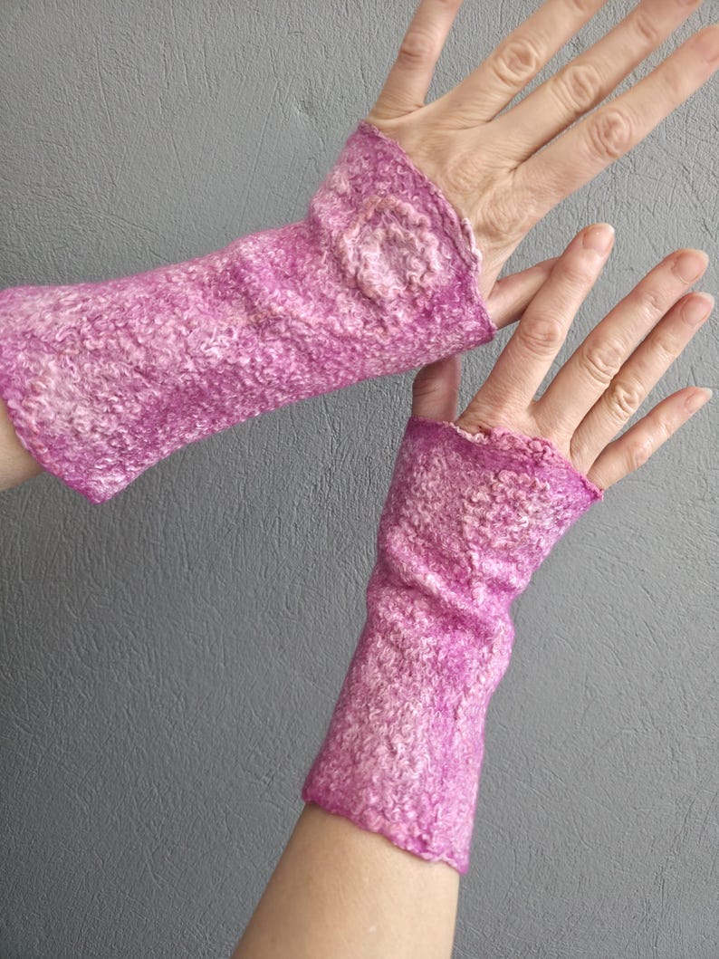 May include: A pair of pink and white felted fingerless gloves. The gloves are a vibrant pink with white flecks, and have a textured, woolly appearance. The wrist cuffs are slightly ruffled. The gloves are designed to keep hands warm while leaving fingers free.